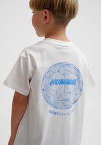 T-shirt blanc à manches courtes avec un graphique bleu d'un ballon de soccer stylisé au dos, comportant le texte "HUMMEL" et "EVERYDAY ACTIVE."