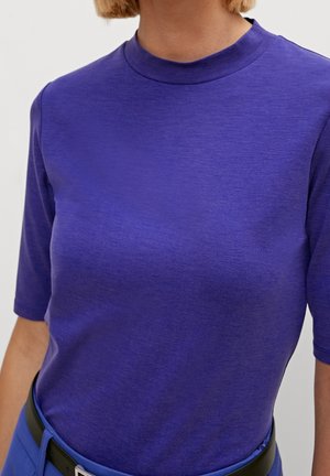 Kvinde iført en tætsiddende lilla kortærmet top, der er stukket ned i blå bukser med et sort bælte, mod en ensfarvet lys baggrund.