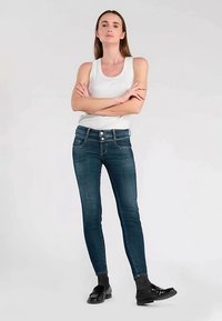 Blauwe skinny jeans met een gladde denimtextuur, voorzien van een klassieke vijfzakkenontwerp en twee knopen aan de voorkant. Gecombineerd met een wit tanktop.