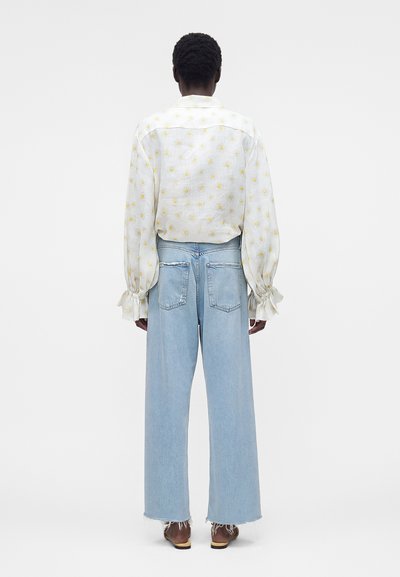 Άτομο με κοντά, σγουρά μαλλιά που φορά λευκό μπλουζάκι με μακριά μανίκια και κίτρινο λουλουδάτο μοτίβο και ανοιχτό μπλε τζιν με φαρδιά μπατζάκια, στέκεται με την πλάτη.