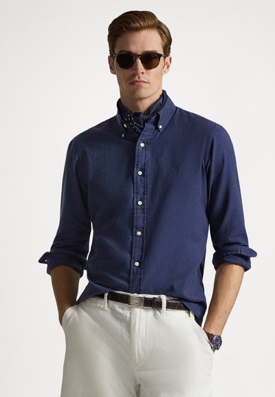 Πουκάμισο σε ναυτικό μπλε με κουμπιά και γυρισμένα μανίκια, λευκό παντελόνι, καφέ ζώνη και γυαλιά ηλίου· διαθέτει μικρό λογότυπο στο στήθος και φουλάρι με σχέδια.