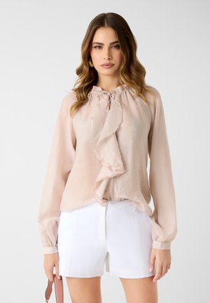 Frau mit welligem braunem Haar, die eine hellrosa Bluse mit langen Ärmeln und Rüschen sowie weiße Shorts trägt und einen Riemen einer braunen Handtasche hält.