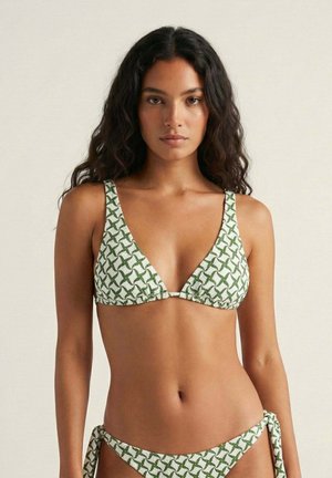 Mujer con cabello largo y rizado, vistiendo un bikini blanco con patrón geométrico verde, de pie sobre un fondo beige claro.