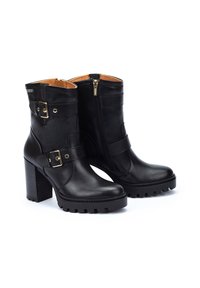 Botas de cuero negro hasta el tobillo con un tacón grueso, que presentan acentos de metal dorado, dos correas ajustables y una cremallera lateral.