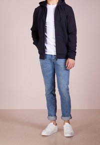 Marinblå zip-up hoodie med dragsko, bärs över en vit T-shirt, i kombination med uppvuxna ljusblå jeans och vita sneakers. Slät textur.