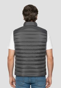 Szara pikowana kamizelka z wysokim kołnierzem, o prążkowanej fakturze i zapinana na zamek, noszona na białej koszuli, prezentująca smukłą sylwetkę.