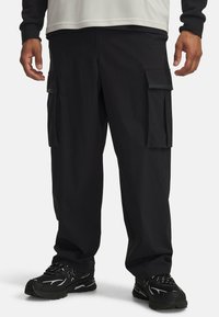 Black cargo pants with a textured fabric. Features multiple side pockets and a relaxed fit, paired with black athletic shoes.