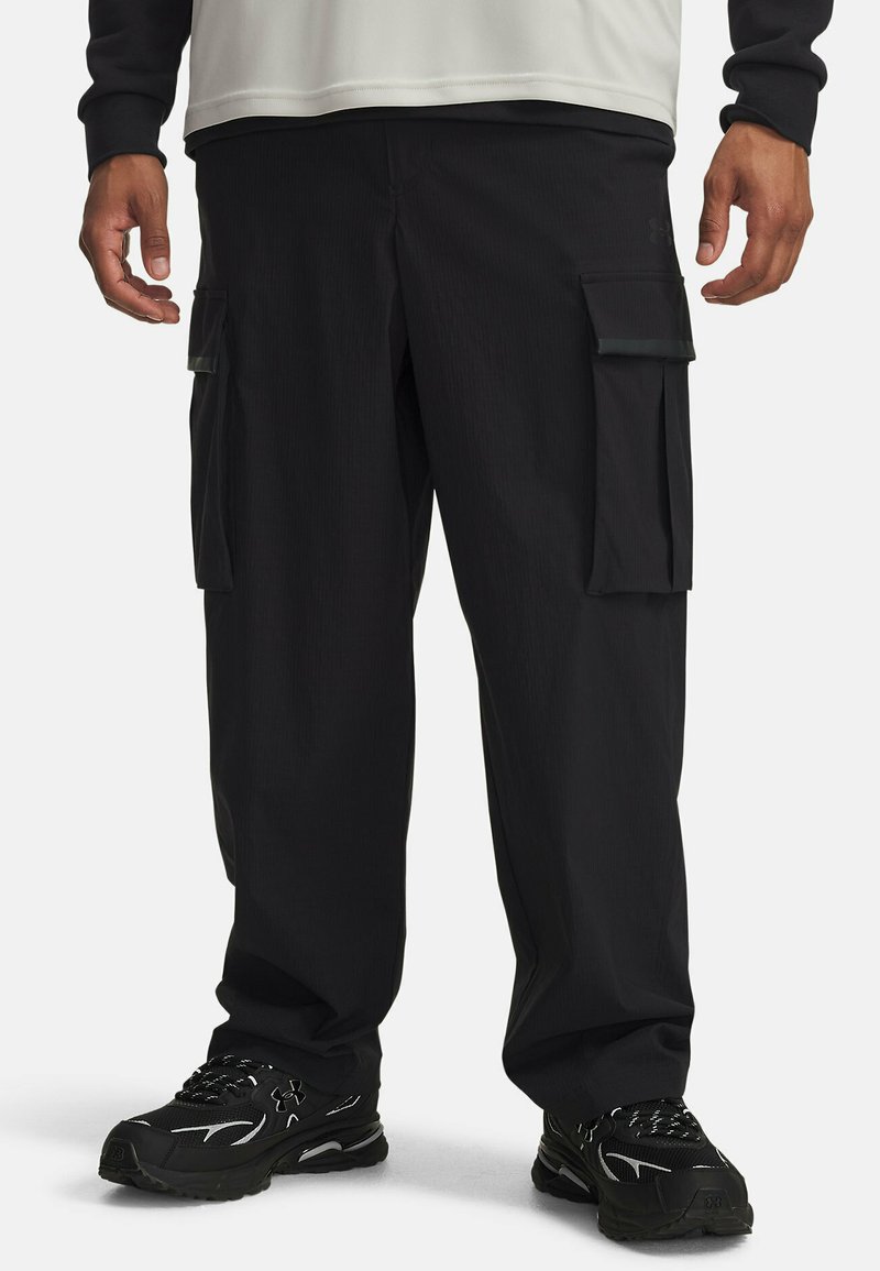 Zwarte cargobroek van een gestructureerde stof. Bevat meerdere zijzakken en heeft een relaxed fit, gecombineerd met zwarte sportschoenen.