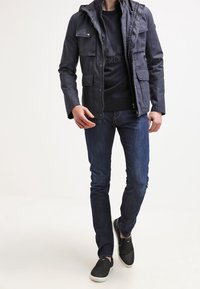 Laivastonsininen takki hupulla ja neljällä etutaskulla, päällä musta neule, yhdistettynä tummansinisiiin farkkuihin ja mustiin liukuvasti pukeutuviiin kenkiin.