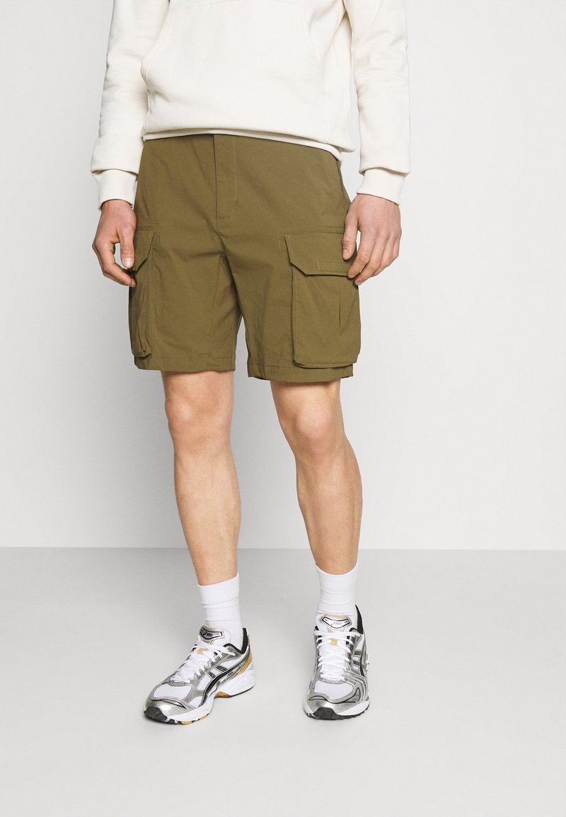 Personne portant un short cargo vert olive, des baskets blanches, des chaussettes blanches et une chemise blanche à manches longues, debout sur un sol gris clair.
