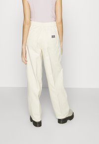 Un pantalon large de couleur crème présente une taille élastique et deux poches arrière avec une petite patch logo. Le tissu semble doux et durable.