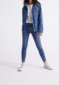 Denimjacka och högmidjade skinny jeans i medelblått. Vit t-shirt under. Vita sneakers. Enkel, figurnära design och slät textur.