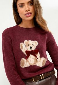 Tumši sarkans džemperis ar pūkainu gaiši brūna tedi lāča dizainu, rotātu ar sarkanu samta loku. Mīksta tekstūra, apaļa kakla izgriezums un garas piedurknes.