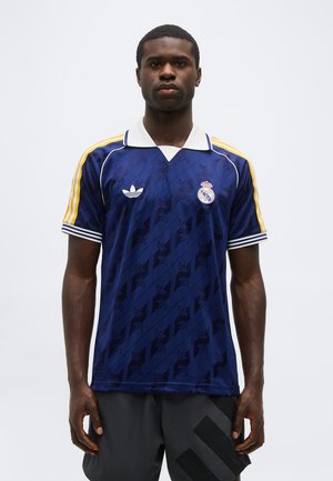 Muž v modrom drese Adidas Real Madrid s bielym golierom a žltými pruhmi na rukávoch stojaci pred jednoliatym bielym pozadím.