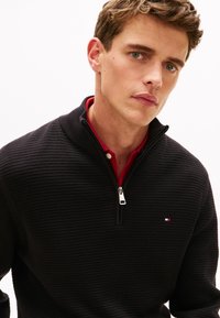 Μαύρο πουλόβερ με φερμουάρ και υφασμάτινο πλεκτό σχέδιο, με μια κόκκινη μπλούζα με γιακά από μέσα και μικρό λογότυπο στο στήθος.