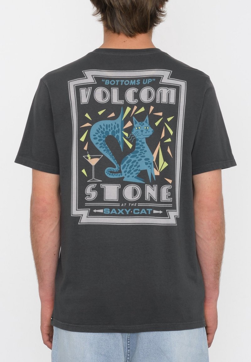 Personne portant un t-shirt sombre avec un graphisme coloré d'un chat bleu tacheté, un cocktail, et le texte "Bottoms Up Volcom Stone Saxy Cat."