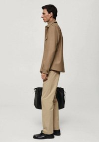 Bruine overhemdd, beige broek, zwarte schoenen, houdend een zwarte tas. De overhemd heeft een kraag en zijzak, en heeft een slim fit silhouet.