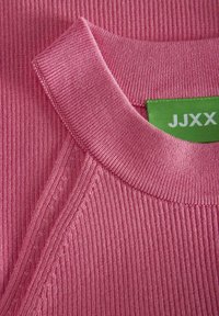 Rožinė giriamasis audinys su plačiu iškirpimu ir žalia etikete, kurioje užrašyta "JJXX". Apima smulkų siuvimą ir glotnią tekstūrą.