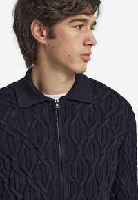 Marinblå stickad zip-up kofta med diamantmönster, ribbad krage och strukturerad design. Har en silverfärgad dragkedja och medeltjock textur.