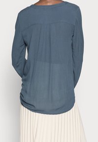 Haut à manches longues bleu ardoise avec un dos froncé, un col rond et une texture lisse, associé à une jupe plissée de couleur claire.