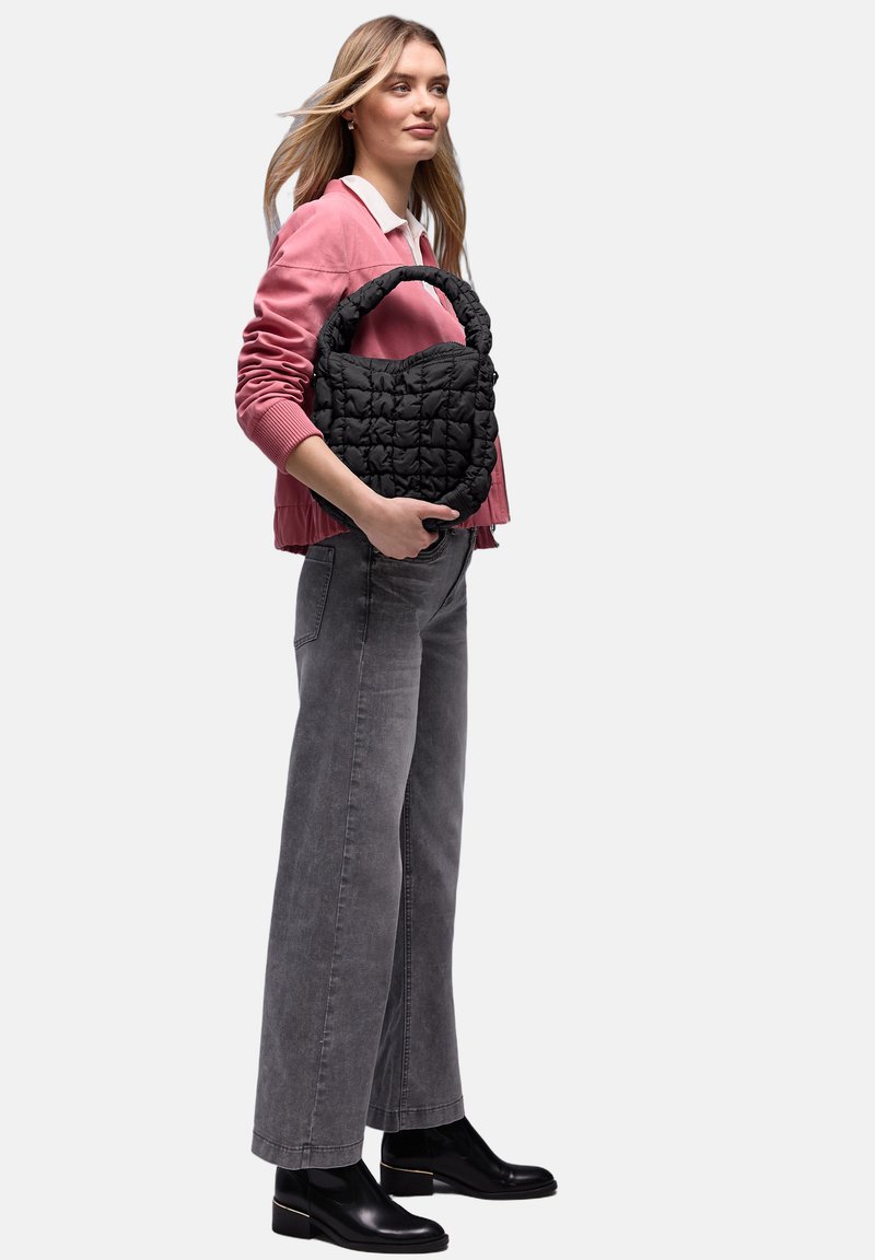 Femme en pull rose et jean large gris tenant un sac à main matelassé noir, portant des bottines noires, debout de profil.