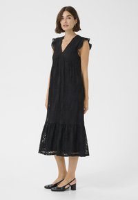 Rochie midi din dantelă neagră, cu mâneci volan, decolteu în V și tiv stratificat. Materialul prezintă modele florale complexe. Purtați cu pantofi cu toc negri.