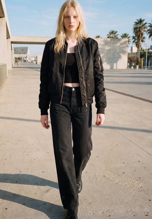 Jeune femme marchant en extérieur vêtue d'une veste bomber noire, d'un crop top noir, d'un jean noir à jambes larges et de chaussures noires sous un ciel dégagé.