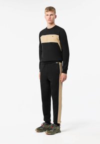 Ensemble de sport noir et beige avec un col rond, des poignets côtelés et des rayures sur les côtés ; comprend un petit logo vert sur la poitrine et sur le pantalon.