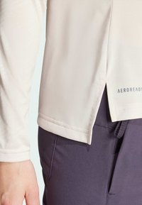 Lichtbeige langemouwen shirt met een gestructureerde stof en zijsplitten, gecombineerd met donkerpaarse broek. "AEROREADY" logo op de zoom van het shirt.