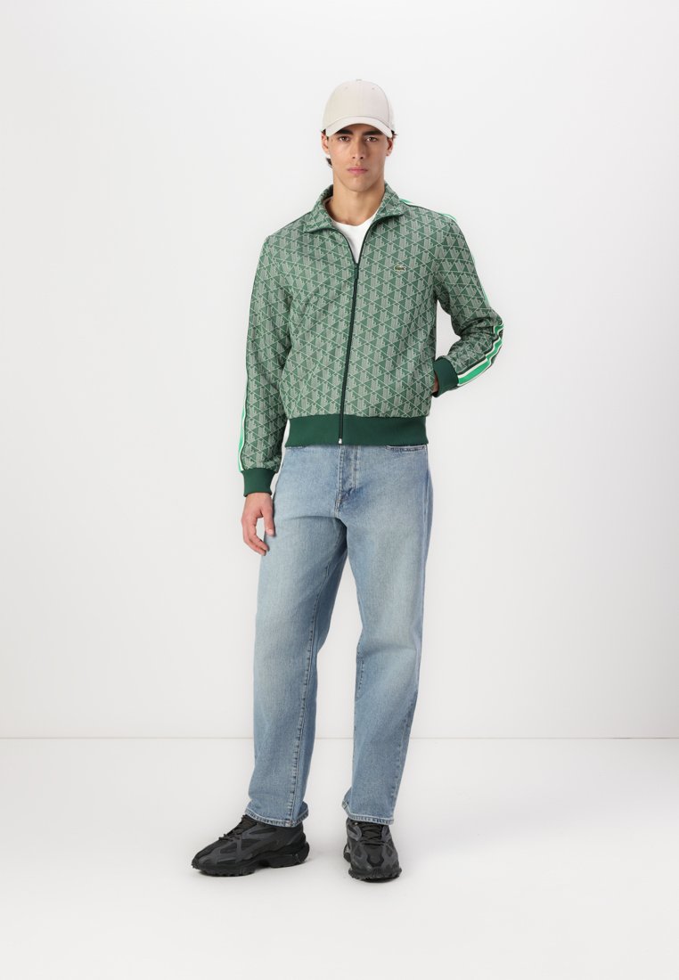 Veste zippée verte avec un motif géométrique, poignets et ourlet à côtes. Associée à un jean bleu clair et des baskets noires. Casquette de couleur claire portée.