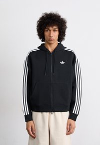 Sweatshirt preto com fecho, feito de algodão, com accentos brancos em três riscas nas mangas e um pequeno logotipo branco no peito. Textura suave.