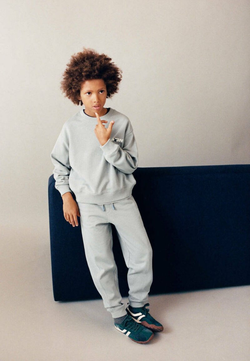 Enfant aux cheveux bouclés portant un sweat-shirt et un pantalon gris, appuyé sur un canapé bleu foncé, touchant ses lèvres pensivement tout en se tenant sur un sol clair.