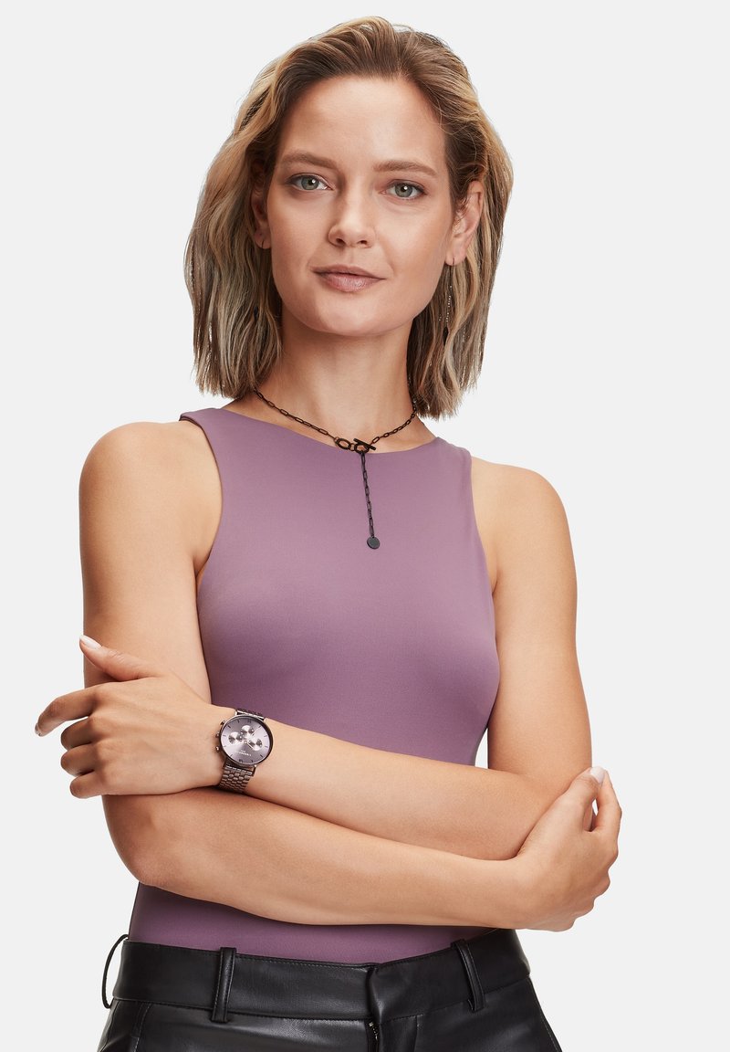 Lila ärmelloses Top aus weichem Stoff, schwarze Perlenkette und eine runde silberne Uhr mit einem netzartigen Armband, kombiniert mit schwarzen Hosen.