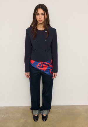 Kobieta o długich brązowych włosach nosi ciemną, dwurzędową marynarkę, asymetryczną bluzkę w niebiesko-czerwone wzory, ciemne podwinięte dżinsy oraz czarne buty.