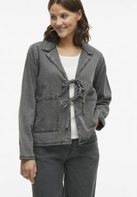 Šedá denimová bunda s uvoľneným strihom, výstrihom do V, uzatváraním na viazanie vpredu a dvoma prednými vreckami. Textúrovaná látka s viditeľnými detailmi šitia.