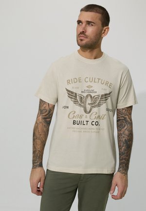 Man with tattooed arms wearing beige "Ride Culture Gas & Grit Built Co." graphic t-shirt and green pants, looking right against plain background.