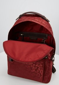 Punainen kangaskeretepack, jossa on hienovarainen leopardikuvio, ruskea nahkainen kantokahva, vetoketjulliset taskut ja käännettävä läppä.