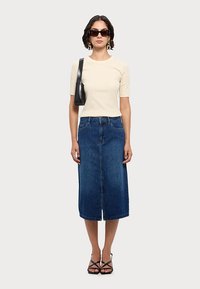 Top bej cu mânecă scurtă, fustă midi din denim albastru închis, geantă de mână neagră și sandale negre cu curele. Modelul poartă ochelari de soare și stă pe un fundal neutru.
