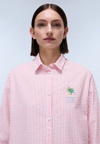 Proužkovaná košile na knoflíky v červené a bílé barvě z bavlny s vyšitým designem palm tree a textem; má špičatý límec a volný střih.