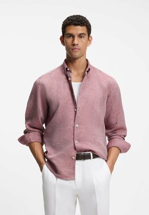 Mann trägt ein hochgekrempeltes langärmeliges pinkes Hemd über einem weißen Unterhemd, weiße Hose und schwarzen Gürtel, die Hände in den Taschen.