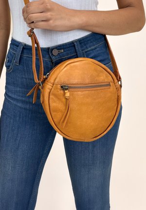Runde, karamelbraune Leder-Crossbody-Tasche mit einer Reißverschlusstasche vorne, verstellbarem Gurt und einer glatten, strukturierten Oberfläche.