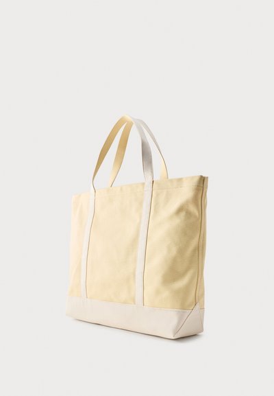Sac fourre-tout rectangulaire beige et crème avec deux bretelles d'épaule, debout sur un fond gris clair uni.