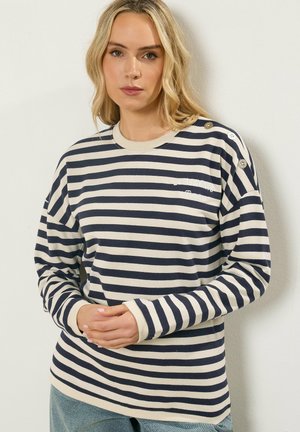 Vrouw met een marineblauw-wit gestreept shirt met lange mouwen en knopen op de schouders, staand tegen een witte muur met handen zachtjes gevouwen.