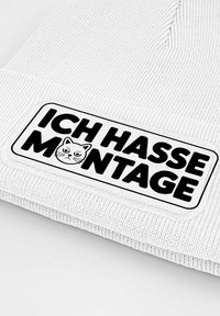 White knit beanie with a textured surface, featuring a rectangular patch that reads "ICH HASSE MONTAGE" with a cat graphic in black.