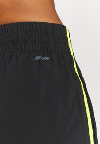 Detailní záběr na pas osoby v černých sportovních šortkách s elastickým pasem a neonově žlutými pruhy po stranách, se značkou "NB DRY".