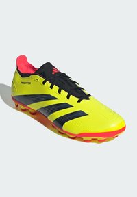 Botín de fútbol amarillo brillante con rayas negras, cordones negros y un interior rojo. Presenta un upper texturizado y una suela robusta con mejor tracción.