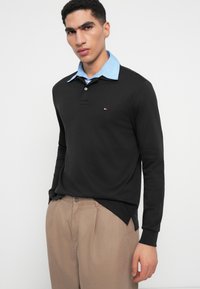 Mladý muž v čiernom polo tričku s dlhým rukávom a modrým golierom stojí proti jednému bielemu pozadiu v béžových nohaviciach.