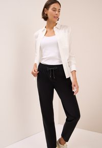 Witte button-up blouse over een witte tanktop, gecombineerd met zwarte broek met een trekkoord in de taille, tegen een neutrale achtergrond.