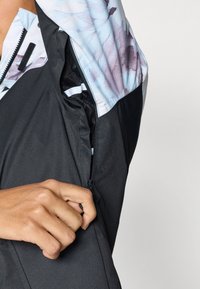 Melna jaka ar ziedu raksta oderējumu un kabatu ar rāvējslēdzēju. Tuvplāns ar roku, kas tur rāvējslēdzēju, izceļot mīksto tekstūru un dizaina detaļas.