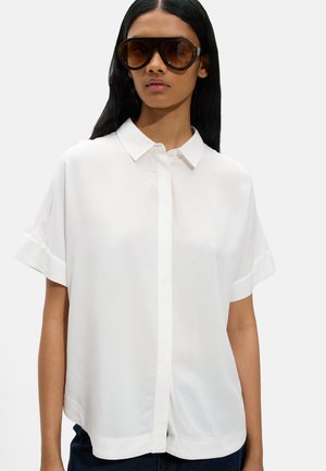 Kobieta w dużych brązowych okularach przeciwsłonecznych i białej koszuli na krótki rękaw z ostrym kołnierzem.